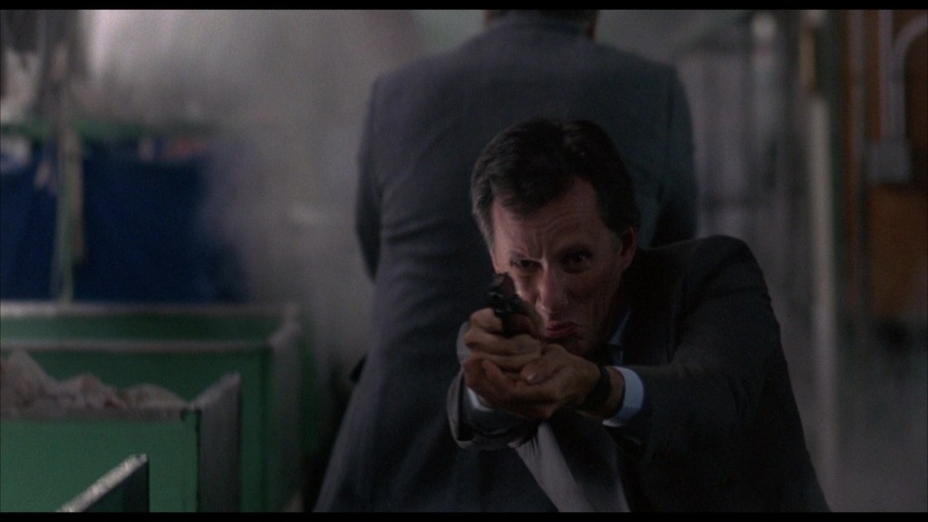 A man in a suit aiming a handgun with a focused expression, set against a blurred background with smoke.