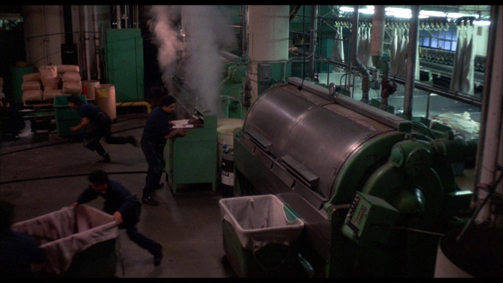A busy industrial setting with workers moving around a large green machine, steam rising from its top. Several bins are present, and large bags are stacked nearby.