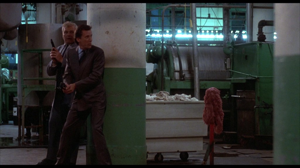 Two men taking cover behind a green and white column in an industrial setting, one holding a firearm and the other looking tense, with machinery visible in the background.