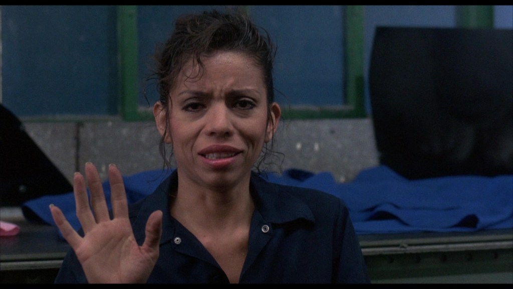 A woman with curly hair, wearing a dark blue outfit, is sitting and raising her hand in a gesture of greeting or acknowledgment, with a concerned expression on her face.
