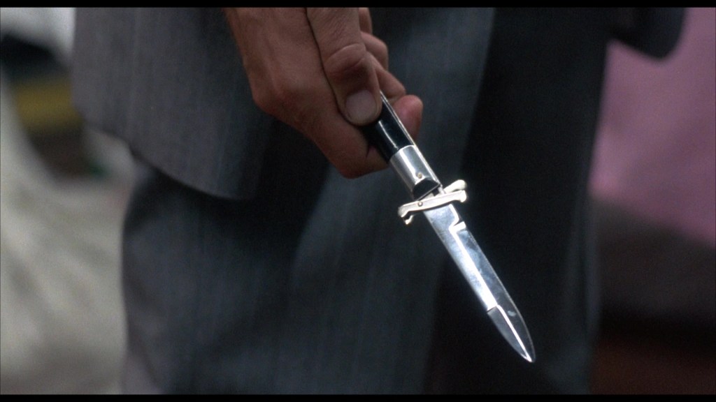 A close-up of a person's hand holding a silver folding knife with a black handle, set against a blurred background.