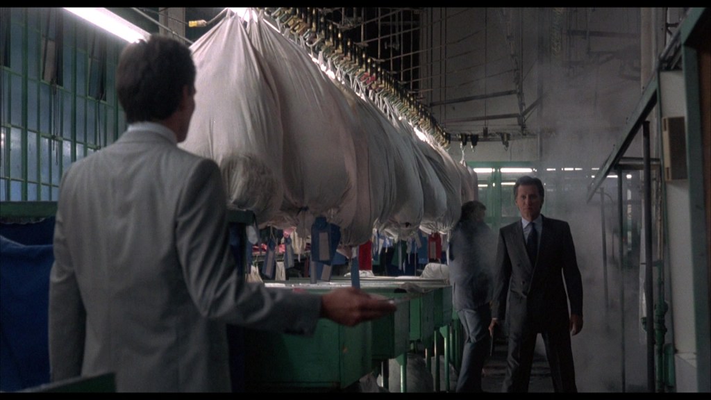 A scene inside a laundromat with hanging garments, featuring two men in suits engaged in conversation. One man stands with his back to the camera while the other faces him, amidst a backdrop of steam and colorful clothing.