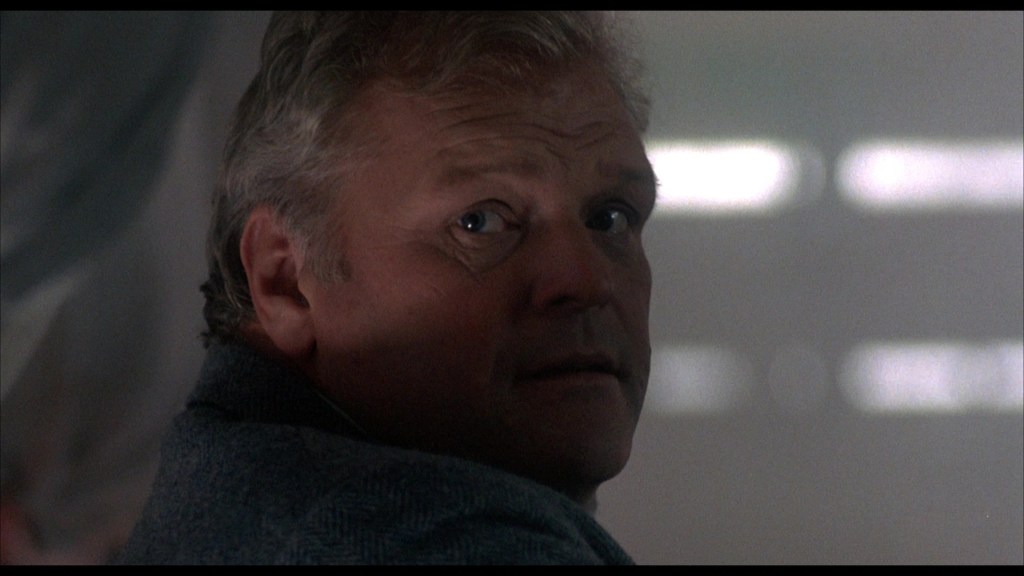 A close-up of a man with short, light-colored hair and a concerned expression, looking over his shoulder.