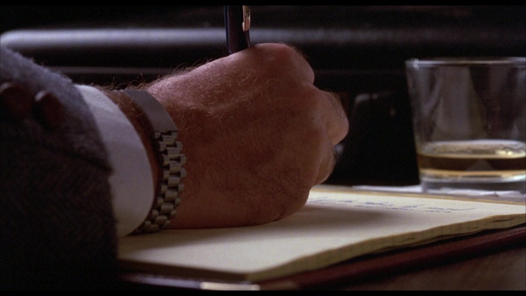 A close-up of a hand writing on a yellow notepad with a pen, accompanied by a glass of whiskey in the background.