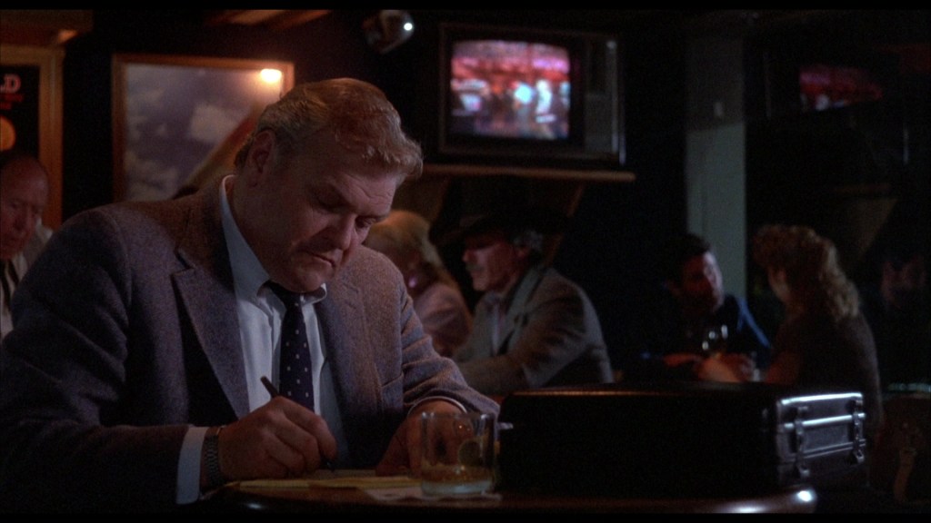 A man in a suit sitting at a bar, writing on a notepad with a drink beside him, while others socialize in the background and a television displays a scene.