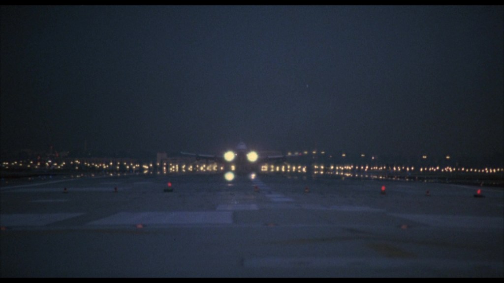 A nighttime airport runway illuminated by lights, with an airplane approaching for landing.