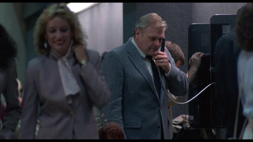 A businessman in a gray suit talking on a payphone at an airport, with people walking by in the background.