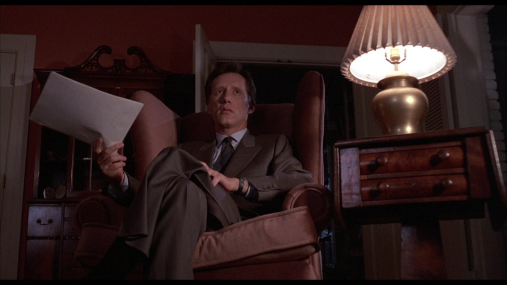 A man in a suit sitting in a large armchair, holding a piece of paper, with a lamp illuminating the scene in a dimly lit room.