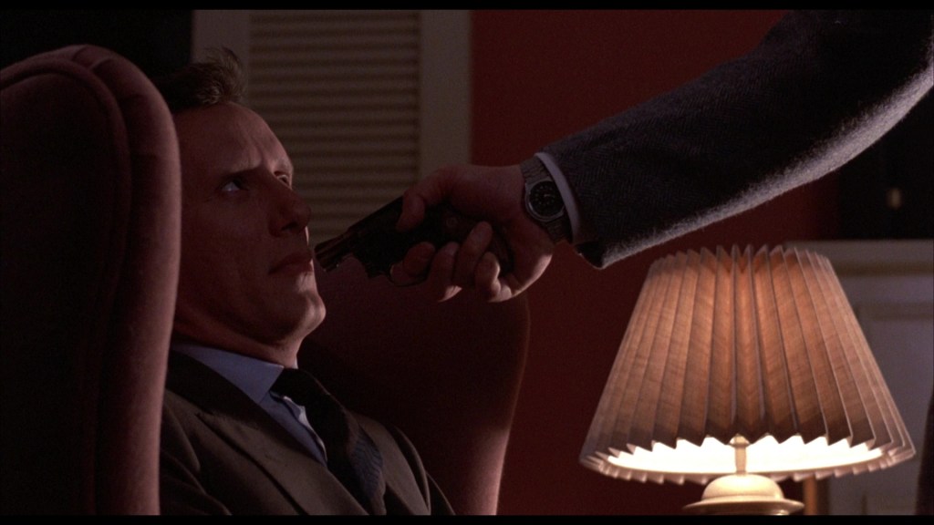 A tense scene where a man sitting in a chair looks fearfully at another man pointing a gun at him. A lamp illuminates the setting, emphasizing the dramatic confrontation.