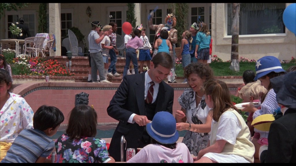 A lively outdoor party scene with children and adults interacting near a swimming pool, decorated with balloons and flowers. In the foreground, two adults, a man in a suit and a woman, are smiling and engaging with a group of children seated around a table.