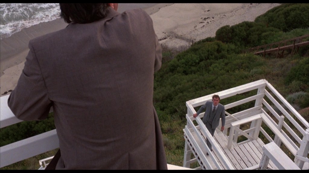 A man in a gray suit stands on a white wooden structure overlooking a beach, while another man in the foreground leans on a railing, observing the scene.