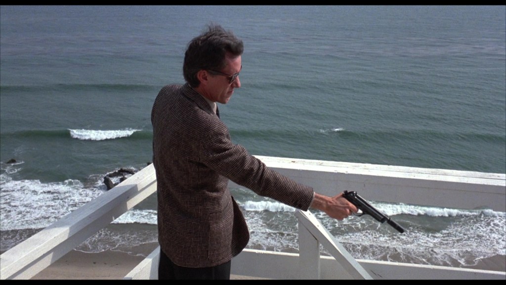 A man in a tweed jacket holds a handgun while standing on a balcony overlooking the ocean and waves.