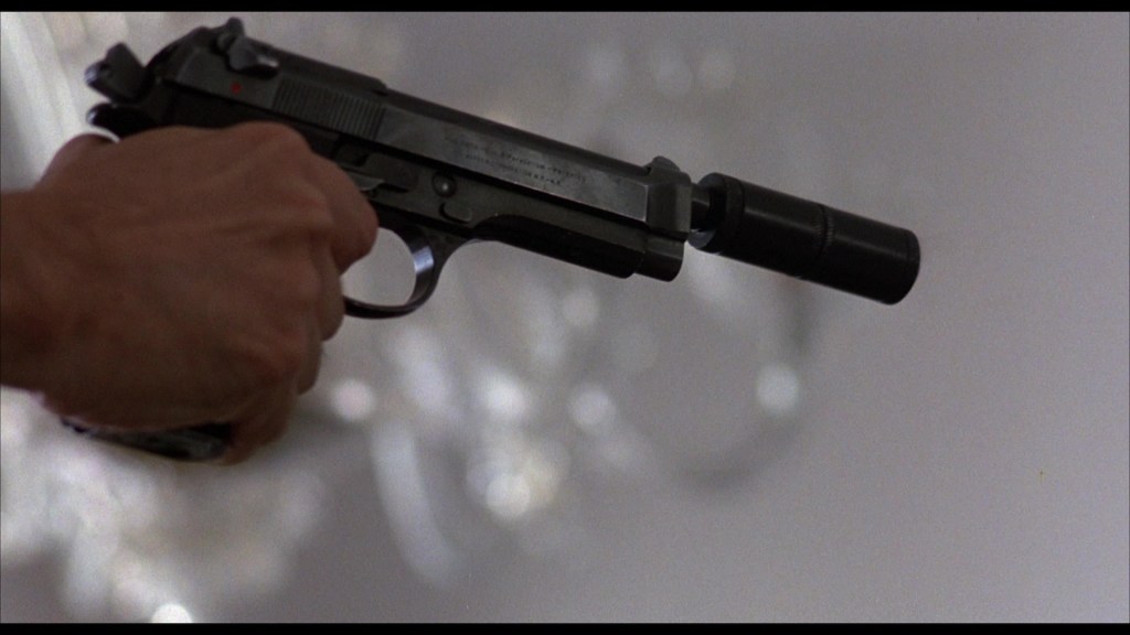 Close-up of a hand holding a handgun with a silencer against a blurred background.