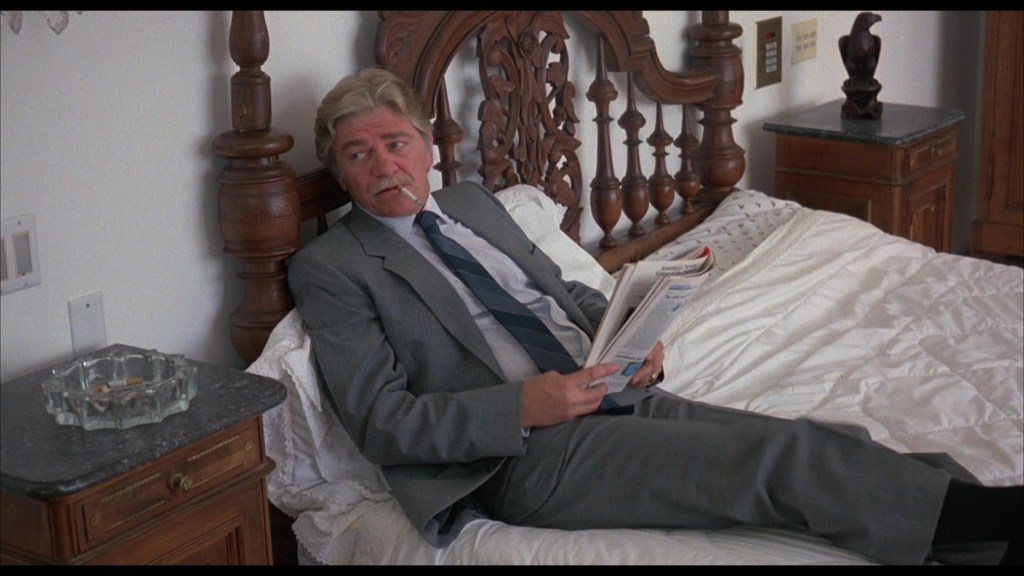 A man in a suit sitting on a bed, reading a newspaper with a cigarette in his mouth. The room features wooden furniture and a nightstand with a glass ashtray.