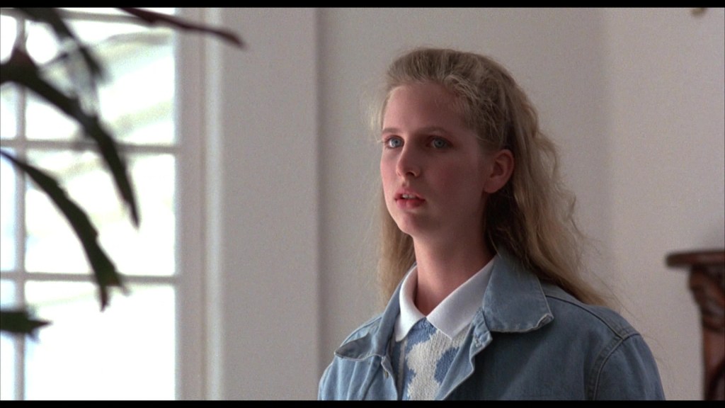 A young woman with long, wavy blonde hair stands in a bright room, looking intently at something off-camera. She is wearing a denim jacket over a white and blue patterned sweater.