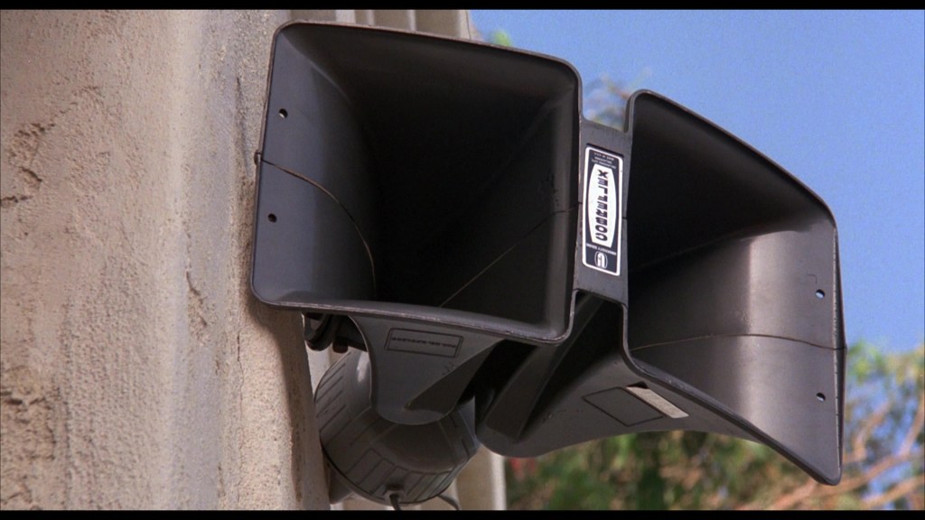 Two large black loudspeakers mounted on a wall, facing outward, with a clear blue sky in the background.