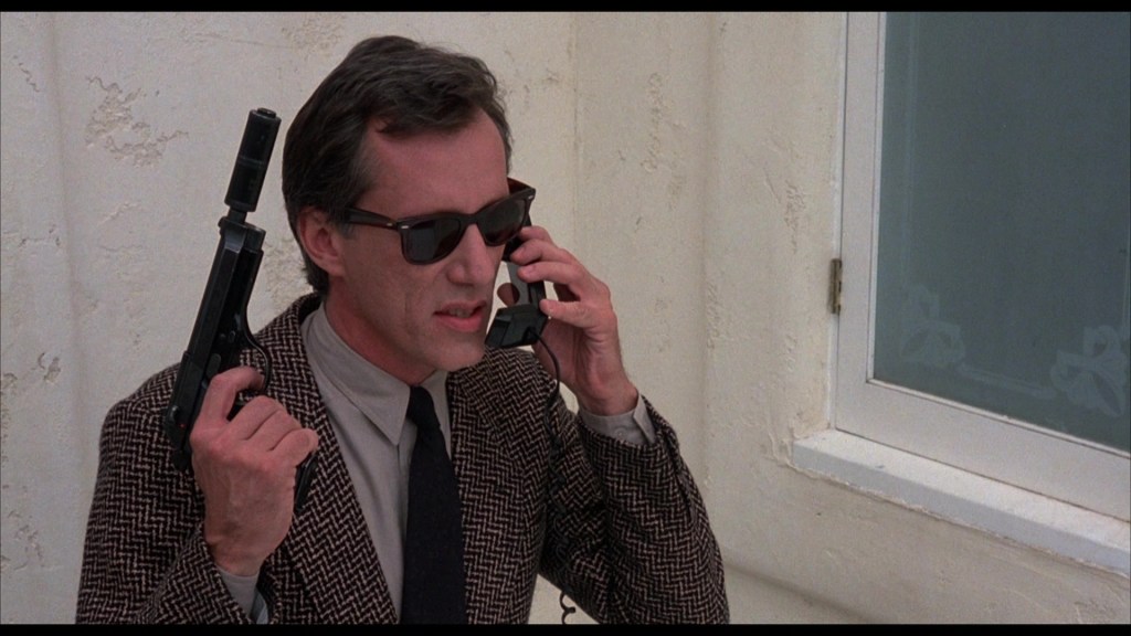 A man wearing sunglasses and a patterned coat is holding a gun in one hand and a phone in the other, standing near a window.