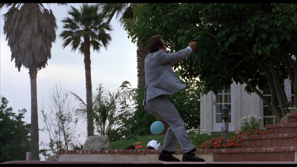 A man in a gray suit stands on a patio with his back turned, holding a gun and appearing to react to a situation. Palm trees and a house can be seen in the background.