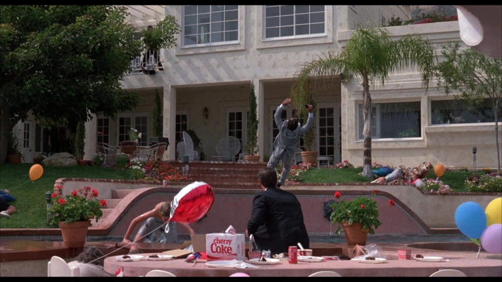 A backyard party scene with colorful balloons, a table with snacks and drinks, and a group of people in formal attire, one person appears to be dancing or celebrating while others are relaxed or lying on the grass.
