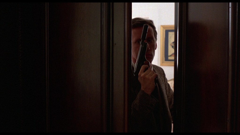 A man holding a gun peering through a partially open wooden door, with a darkened background.