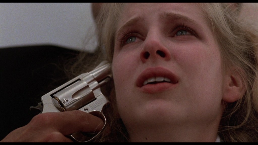 A close-up of a distressed young woman with tears in her eyes, looking upwards with a fearful expression as a hand aims a silver revolver at her head.