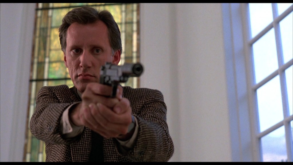 A man in a checkered blazer aiming a handgun in an indoor setting with stained glass windows in the background.