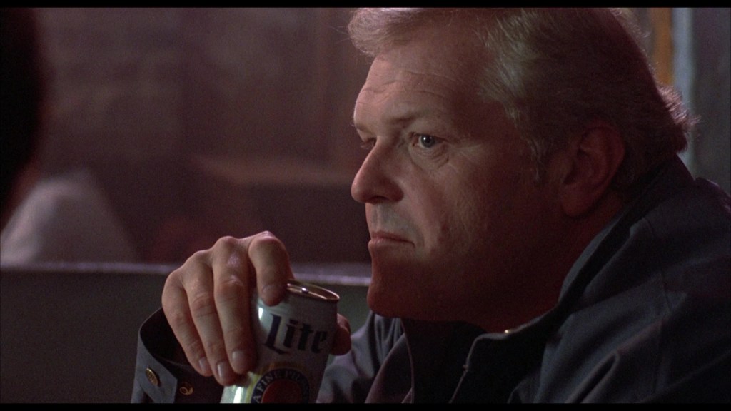 A close-up of a man with light hair, sitting thoughtfully and holding a can of Lite beer, in a dimly lit setting.