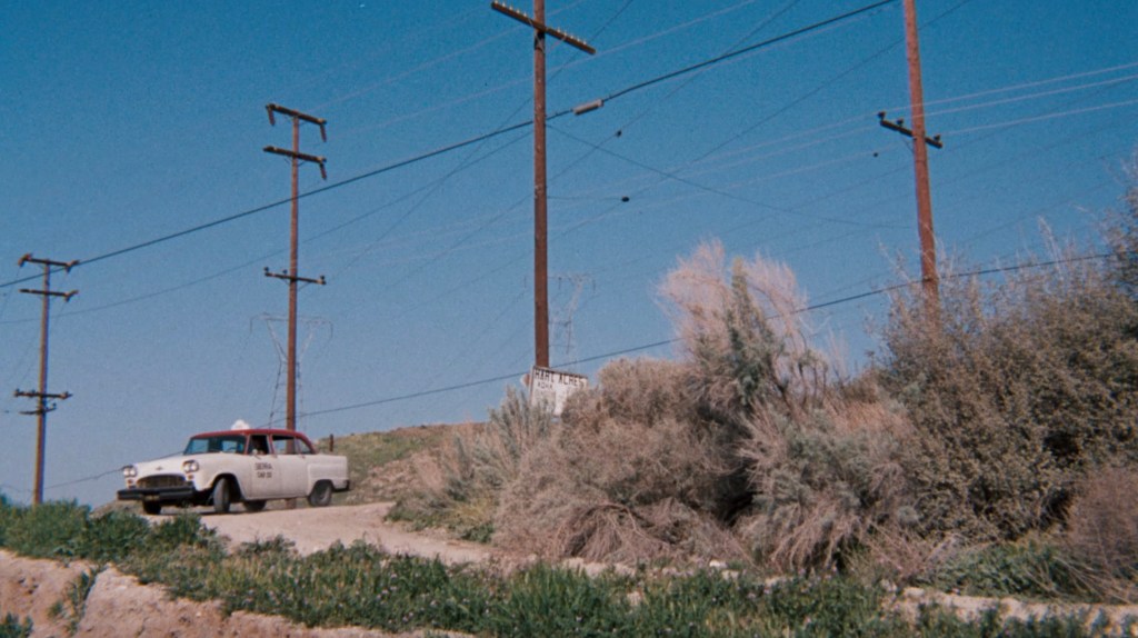 A vintage white taxi parked on a dirt road with utility poles and blue sky in the background