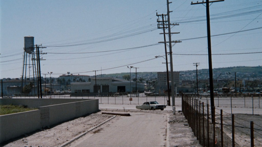 A wide view of an empty lot with a dirt road leading into the foreground, surrounded by a fence and power lines. In the distance, a water tower and a building are visible under a clear sky.