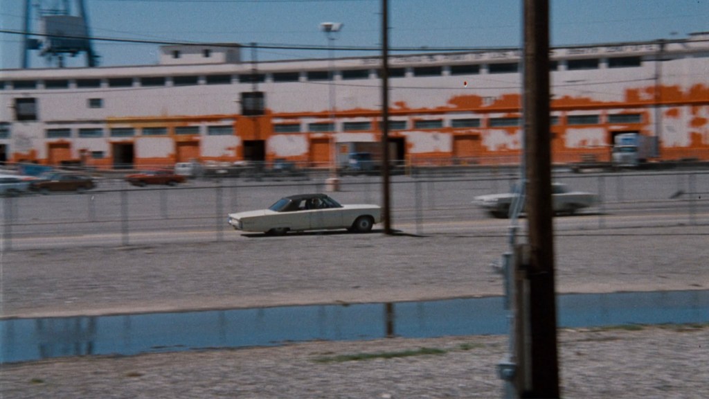 A white car drives along a gravel lot near a large, partially painted building. Power lines and a water reflection are visible in the foreground.