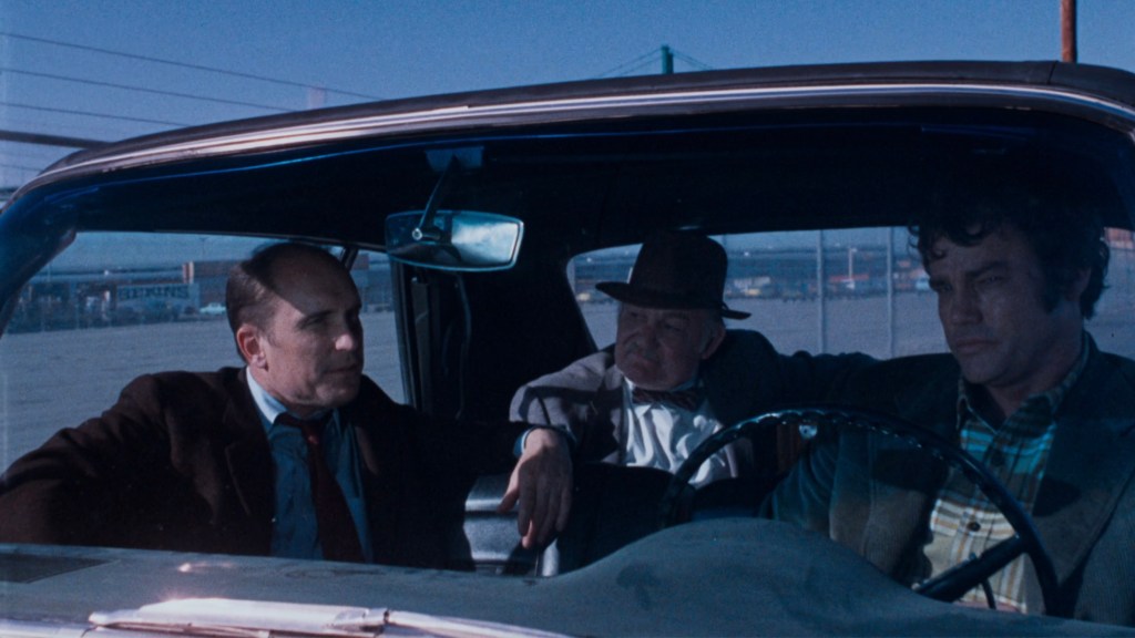 Three men sitting in a car, with the Bay Bridge visible in the background.