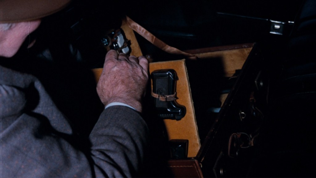 Close-up of a person's hand reaching into a brown suitcase, revealing a black object inside, possibly a firearm.