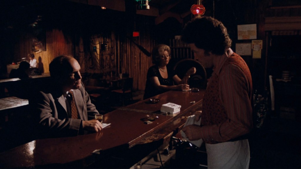 A dimly lit bar scene with a man in a suit at the counter, a woman in a black dress seated nearby, and a bartender wearing a red shirt and apron attending to them.