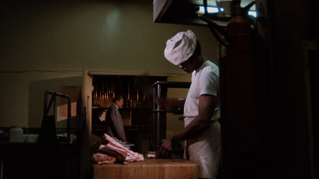 A chef wearing a white shirt and hat is preparing food on a wooden counter while a man in a suit stands in the background.