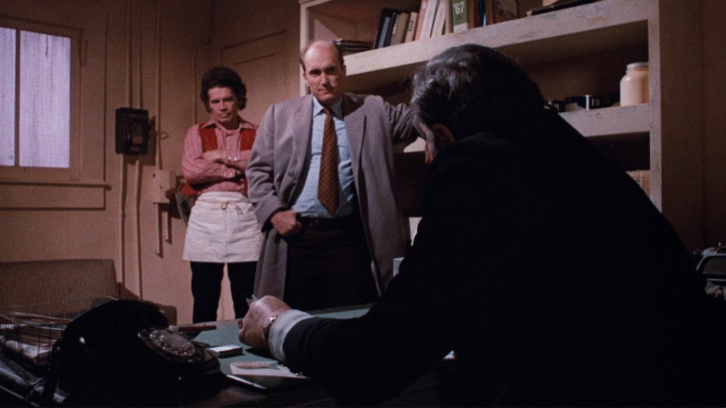 A tense scene featuring three characters in an office setting. One character stands with arms crossed, another sits with a notepad, and a third leans against a shelf, all engaged in conversation.