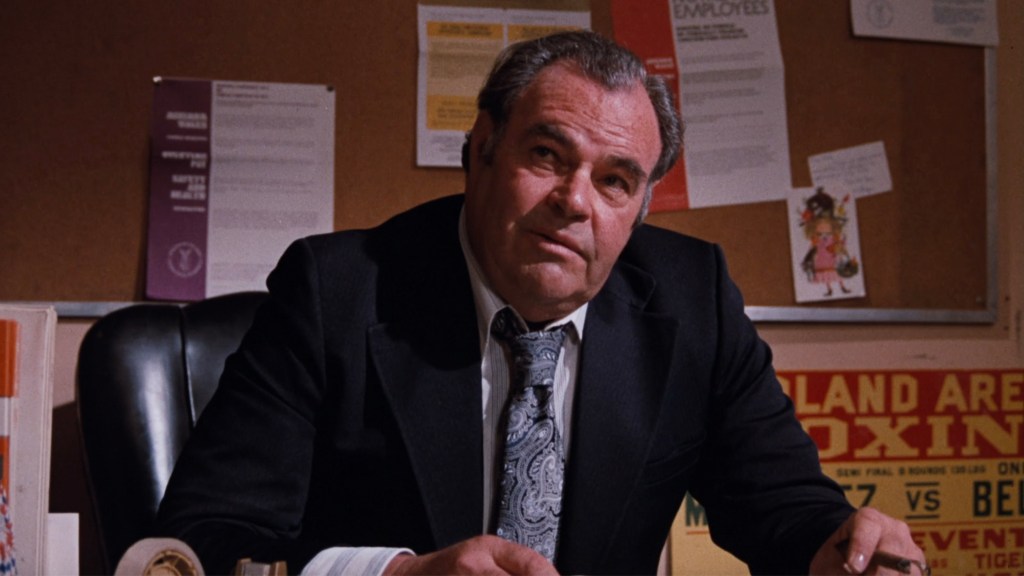 A middle-aged man in a suit sits at a desk, looking thoughtfully to the side with a serious expression. The background features a corkboard with various papers and notices.