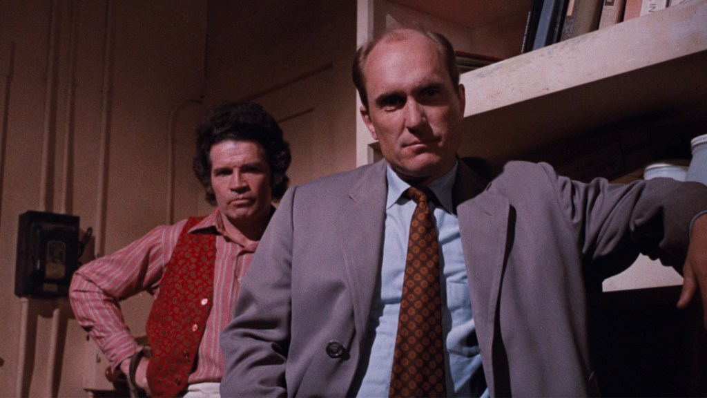 Two men in a room, one wearing a gray suit with a patterned tie, looking serious, while the other, in a red and white striped shirt with a vest, stands behind him, appearing concerned.