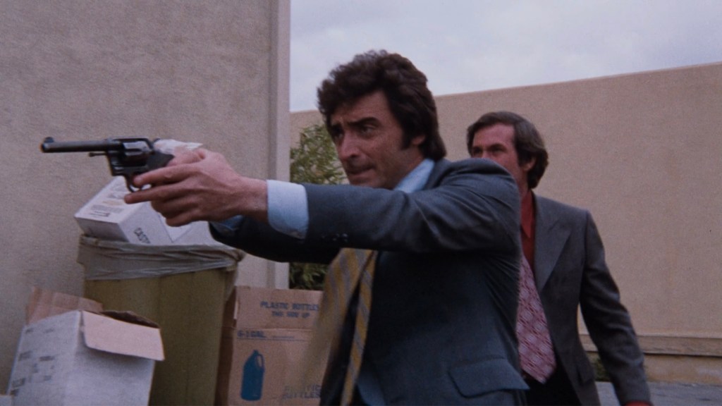 A man in a suit aiming a revolver while another man follows closely behind, set against a backdrop of urban debris.