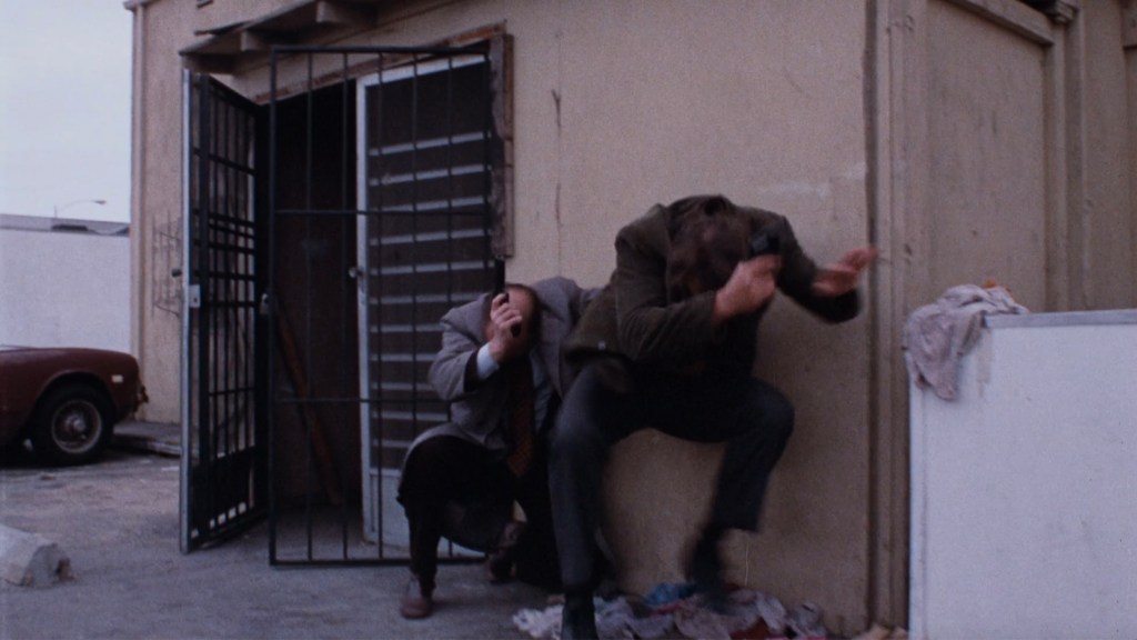 Two men taking cover behind a wall during a tense standoff, one holding a gun and the other protecting his face, with an open barred door in the background.