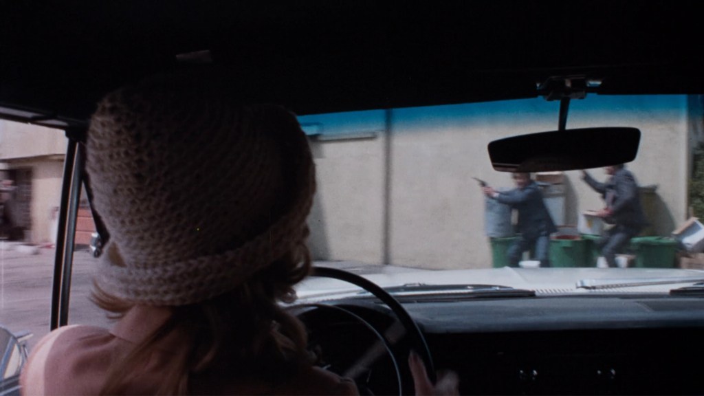 A driver with a crochet hat looking out the car window while two men outside point guns at each other.