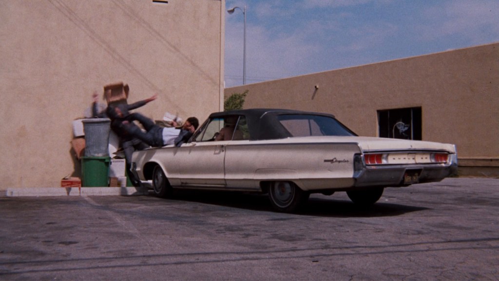 A vintage car parked next to a dumpster, with two people falling out of the car and onto a pile of boxes.