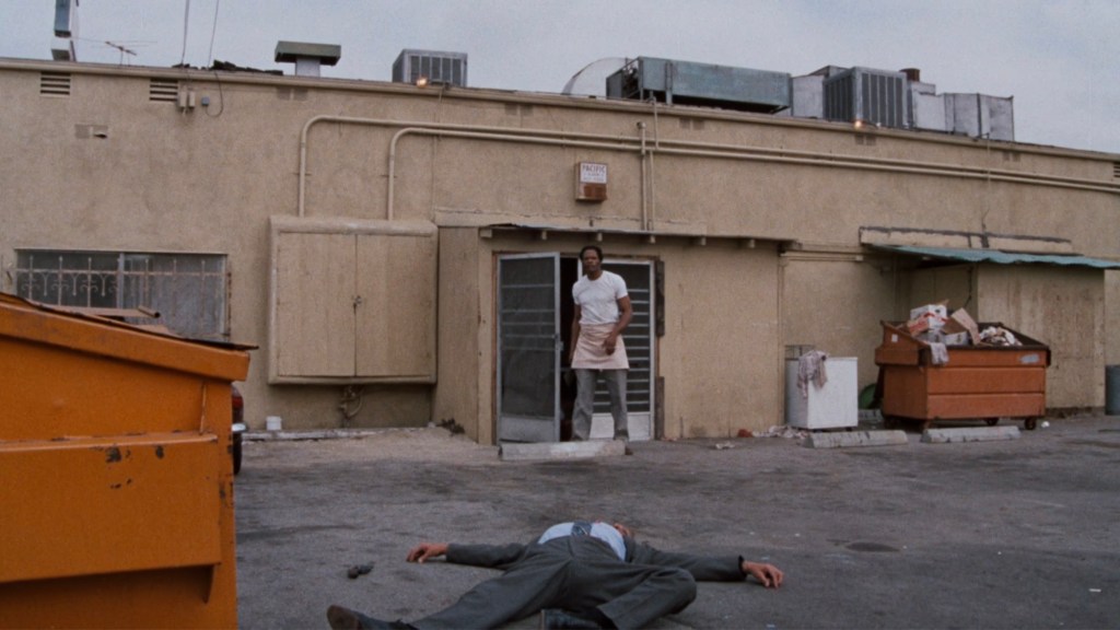 A man in a white shirt and apron stands in an alleyway next to a doorway, looking at a body lying on the ground. In the background, there are dumpsters and a beige building.