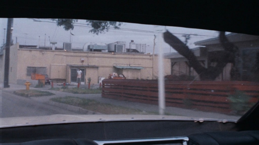 View of a street scene through a car windshield, featuring a beige building, a green awning, a tree, and a red wooden fence.