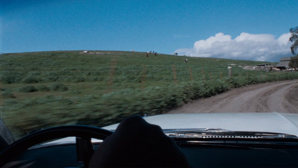 View from inside a car driving along a dirt road, with green fields and a blue sky visible in the distance. A hand is seen on the steering wheel, and there are distant figures walking on the hill.