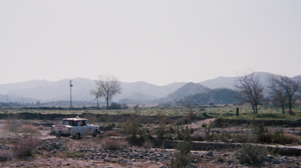 A white car driving through a rocky, grassy landscape with mountains in the background and a clear sky.