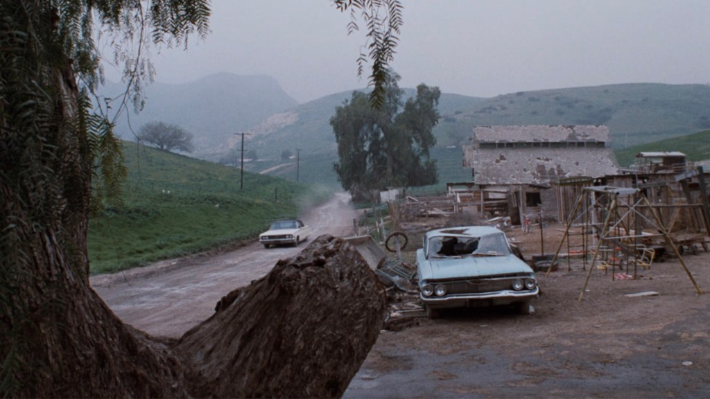 A rural landscape with a dirt road, two parked cars, and an abandoned building in the background. Rolling hills and trees surround the area under a cloudy sky.