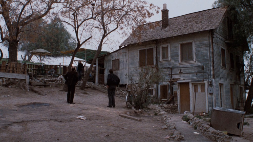 Two figures in dark clothing standing on a dirt path in front of a dilapidated wooden house surrounded by bare trees.
