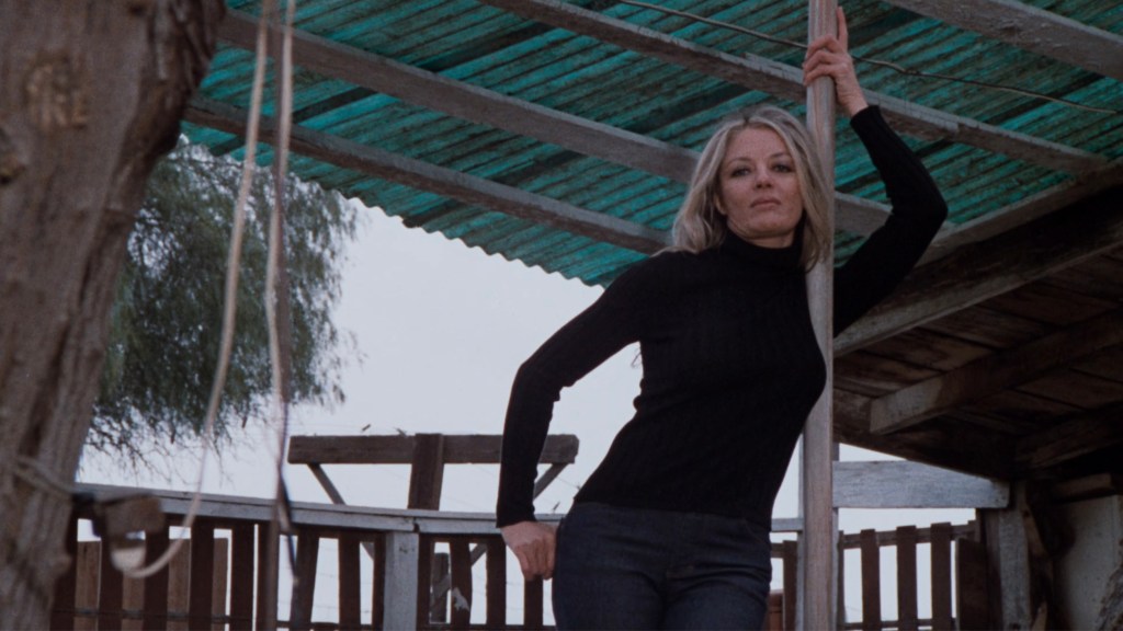 A woman in a black turtleneck leans against a wooden pole under a teal roof, with a rustic setting in the background.