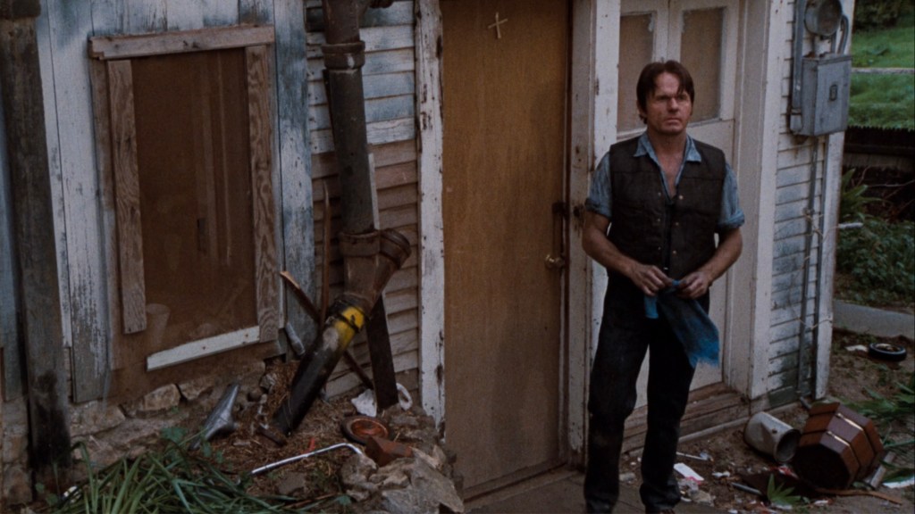 A man stands outside a run-down house, holding a blue cloth, with a worn door behind him and various debris scattered at his feet.