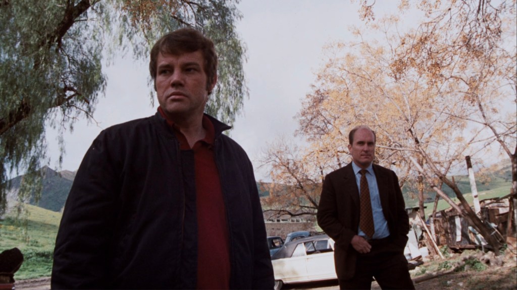 Two men stand outdoors against a backdrop of trees and hills, one casually dressed and the other in a suit, highlighting a serious moment.